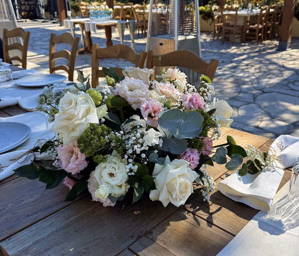 Floral centerpiece with white and pink roses and greenery on a wooden table set for outdoor dining.