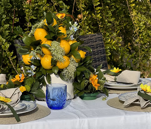 Outdoor dining table set with beige placemats, floral plates, linen napkins with yellow flowers, and a centerpiece of lemons and green foliage in a white vase.