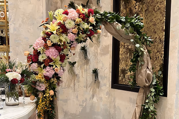 Decorative floral arch with colorful flowers and green leaves draped with beige fabric in an indoor setting.