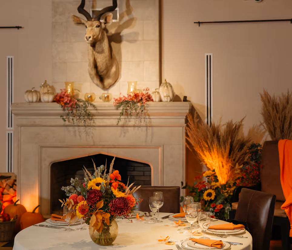 Cozy autumn dining table with floral centerpiece, orange napkins, and a mounted deer head above the fireplace.
