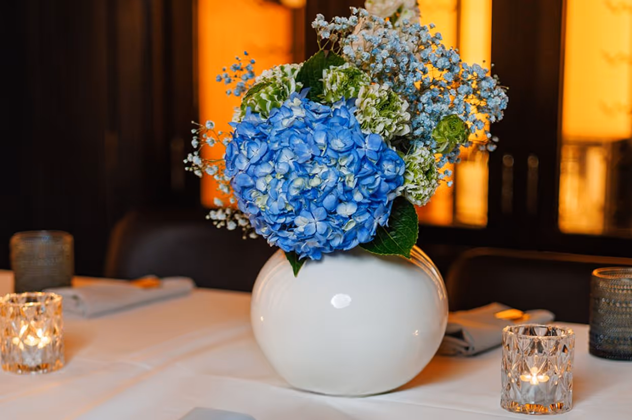 Blue and green flower arrangement in white vase on a table with lit candles and folded napkins.