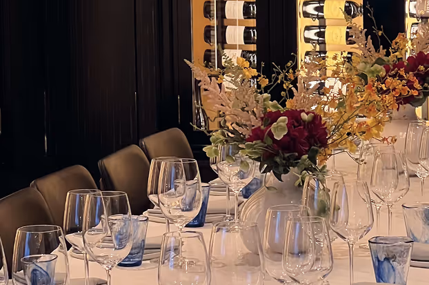 Elegant dining table set with wine glasses, blue water glasses, and a vase of mixed flowers in a sophisticated room with wine bottles displayed in the background.