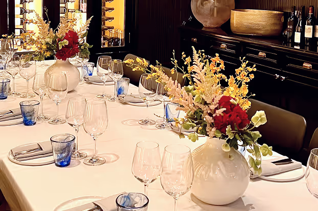 Elegant dining table set with white tablecloth, wine glasses, blue water glasses, silverware, and white vases holding colorful floral arrangements.