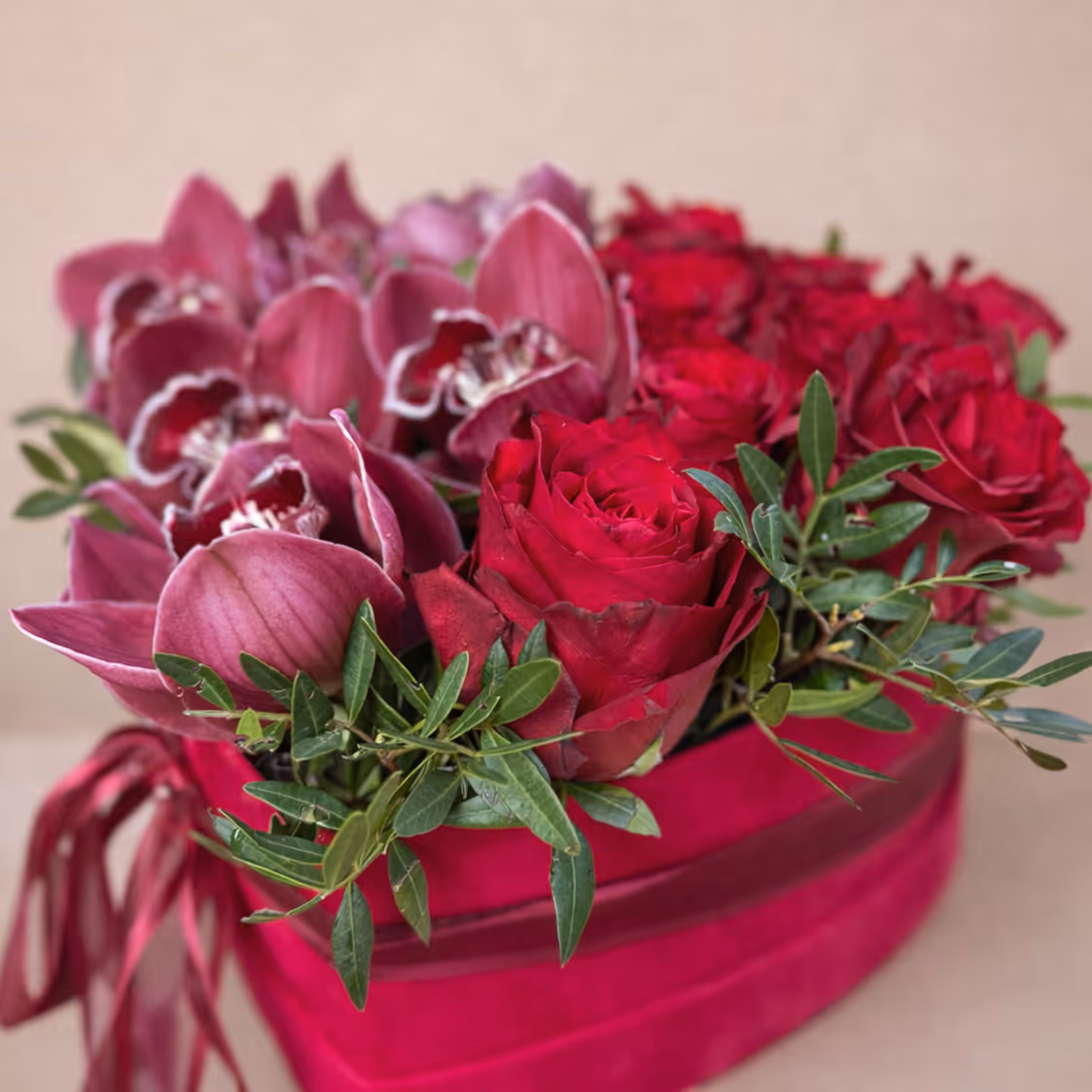 Red roses and purple orchids arranged with green leaves in a pink decorative basket.