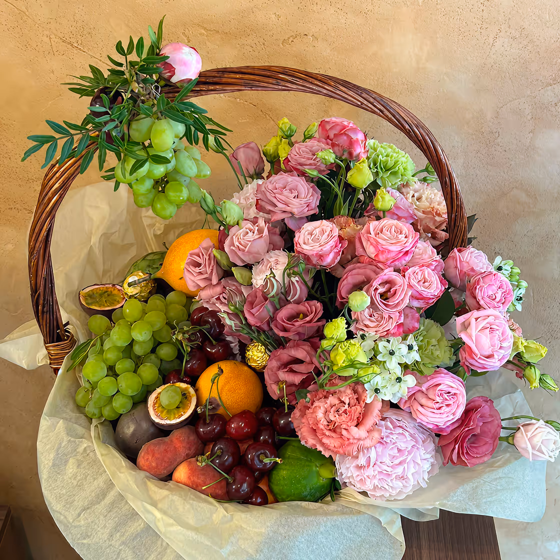 Wicker basket filled with pink flowers and assorted fresh fruits including grapes, oranges, and cherries.