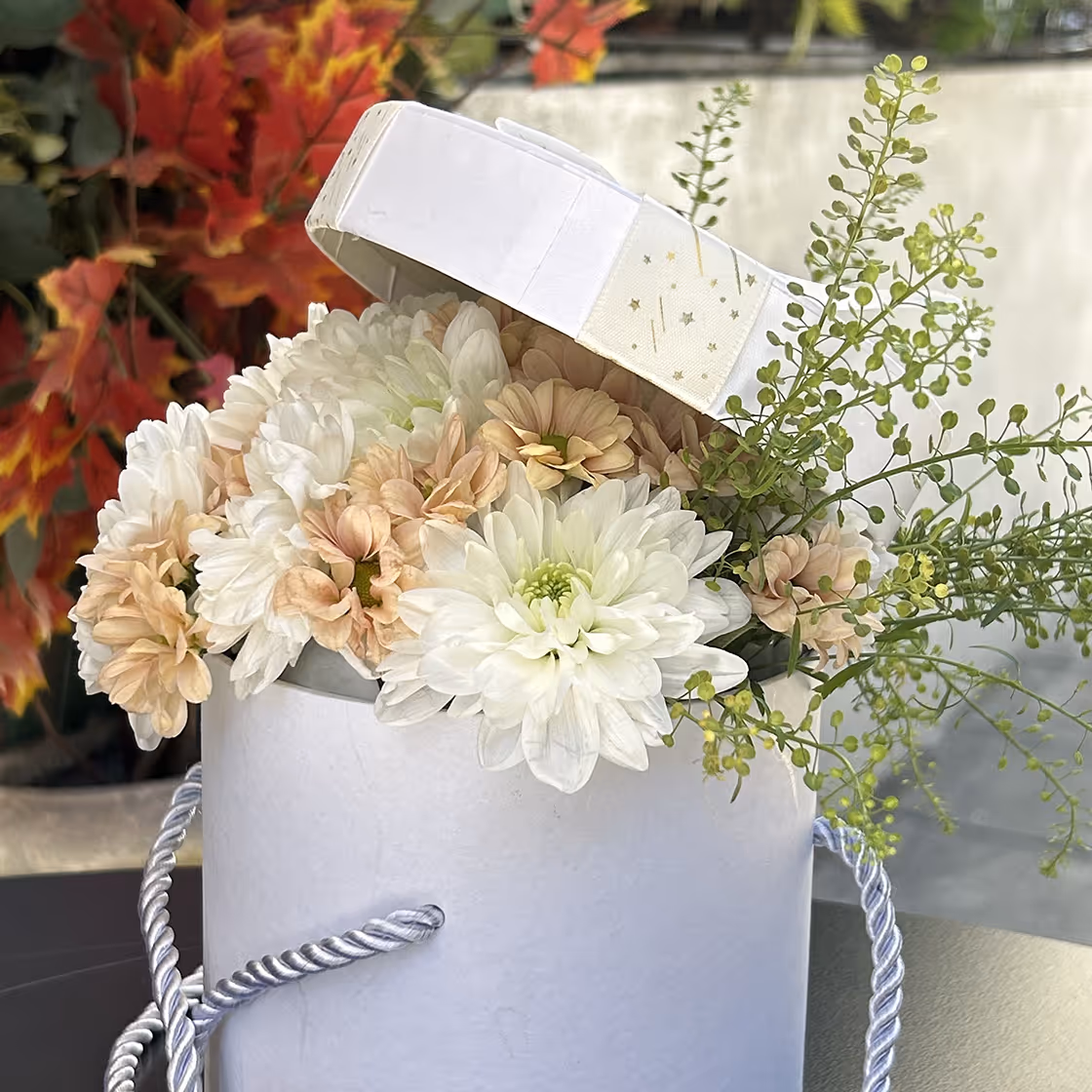 White hatbox with lid angled open showing bouquet of white and pale peach flowers and green foliage.