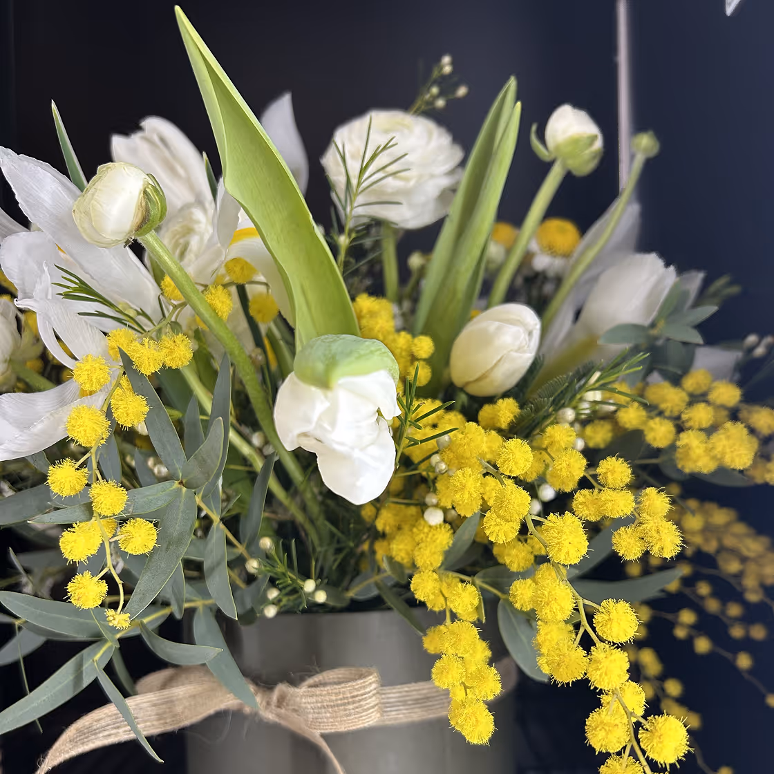Bouquet of white flowers and bright yellow mimosa blooms with green leaves in a tied vase.