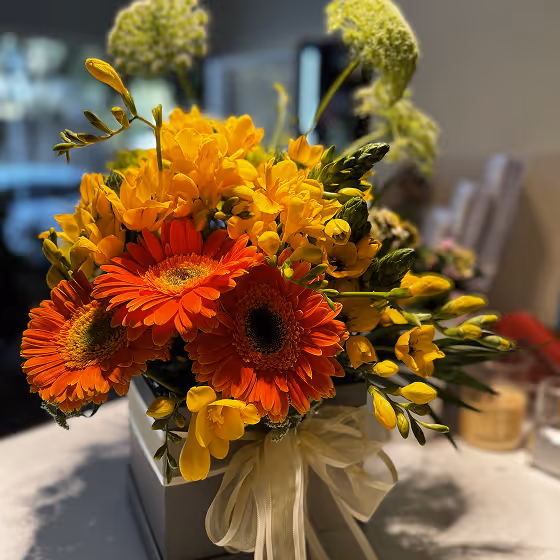 Bouquet of vibrant orange and yellow flowers in a white box with a sheer white ribbon.