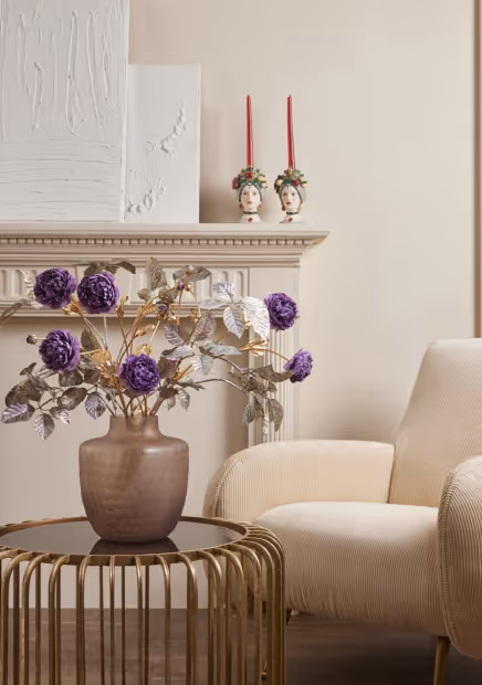 Indoor scene with a beige armchair, a gold metal side table holding a brown vase of purple flowers, and a fireplace mantel with two decorative candle holders.