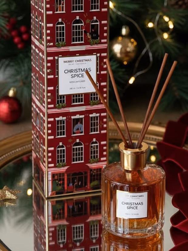 Christmas Spice scented reed diffuser in a faceted glass bottle with gold cap, standing next to its red decorative box shaped like a building, with holiday decorations in the background.