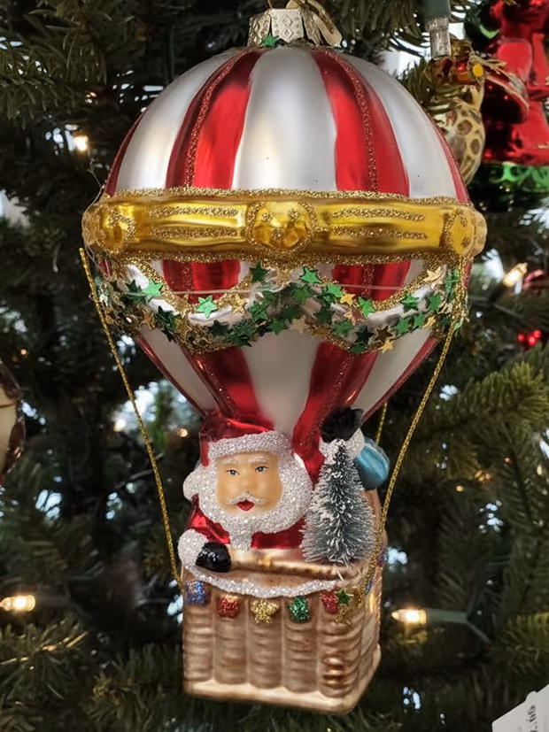 Christmas tree ornament of Santa Claus in a hot air balloon decorated with red and white stripes, gold trim, and green stars, holding a small Christmas tree.