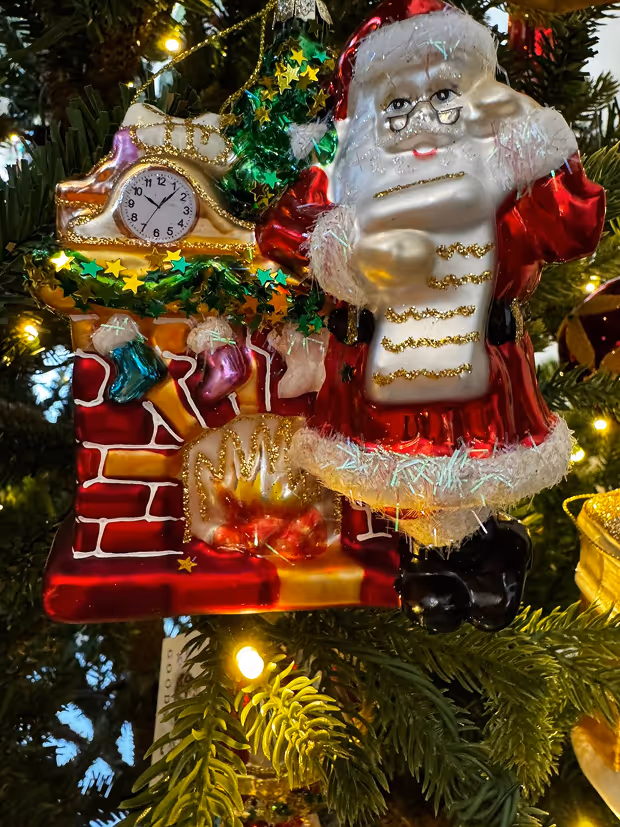 Christmas tree ornament featuring Santa Claus with a list standing beside a decorated fireplace with stockings and a clock.
