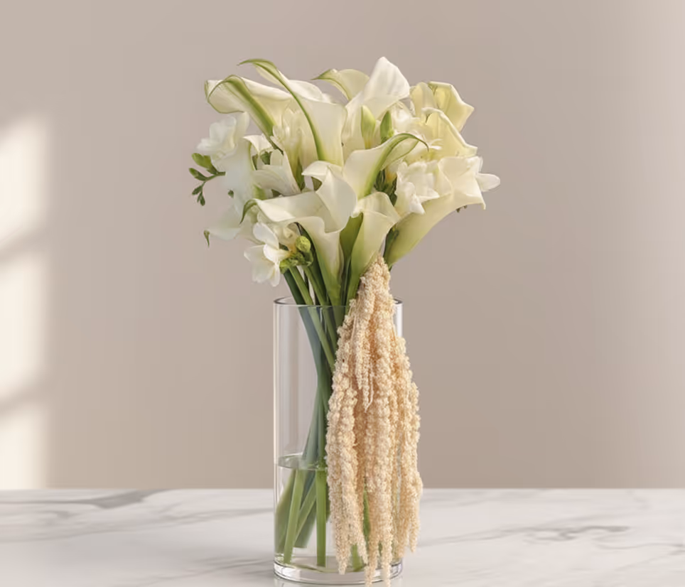 Glass vase with white calla lilies and hanging beige dried flowers on a marble surface.