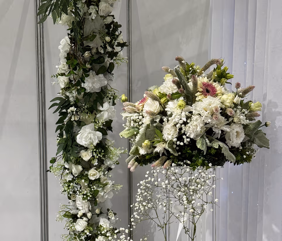 White floral arrangement with greenery on a stand beside a hanging floral garland on a gray wall.