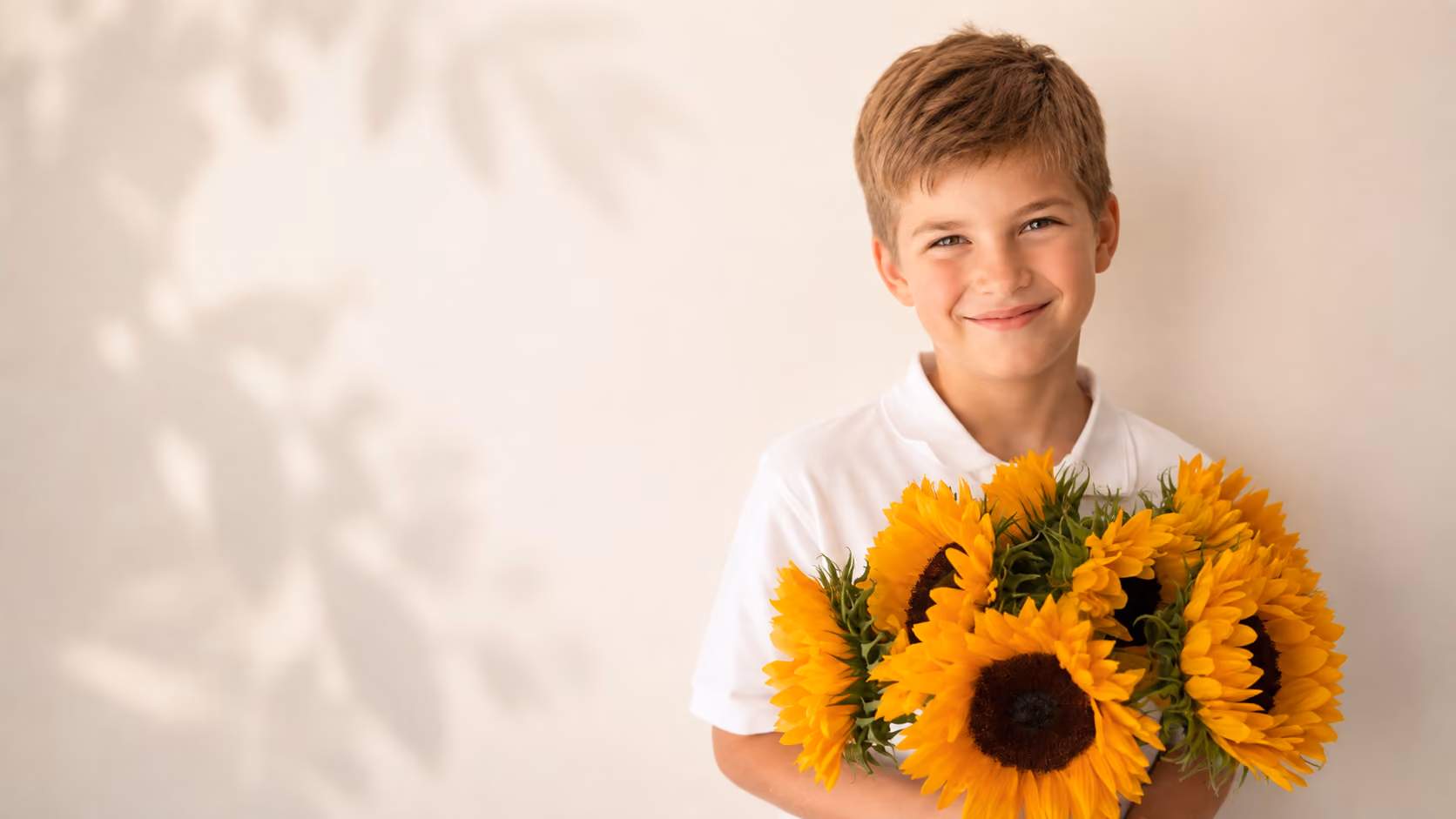 Smiling boy in white shirt holding a bouquet of bright yellow sunflowers against a beige background.