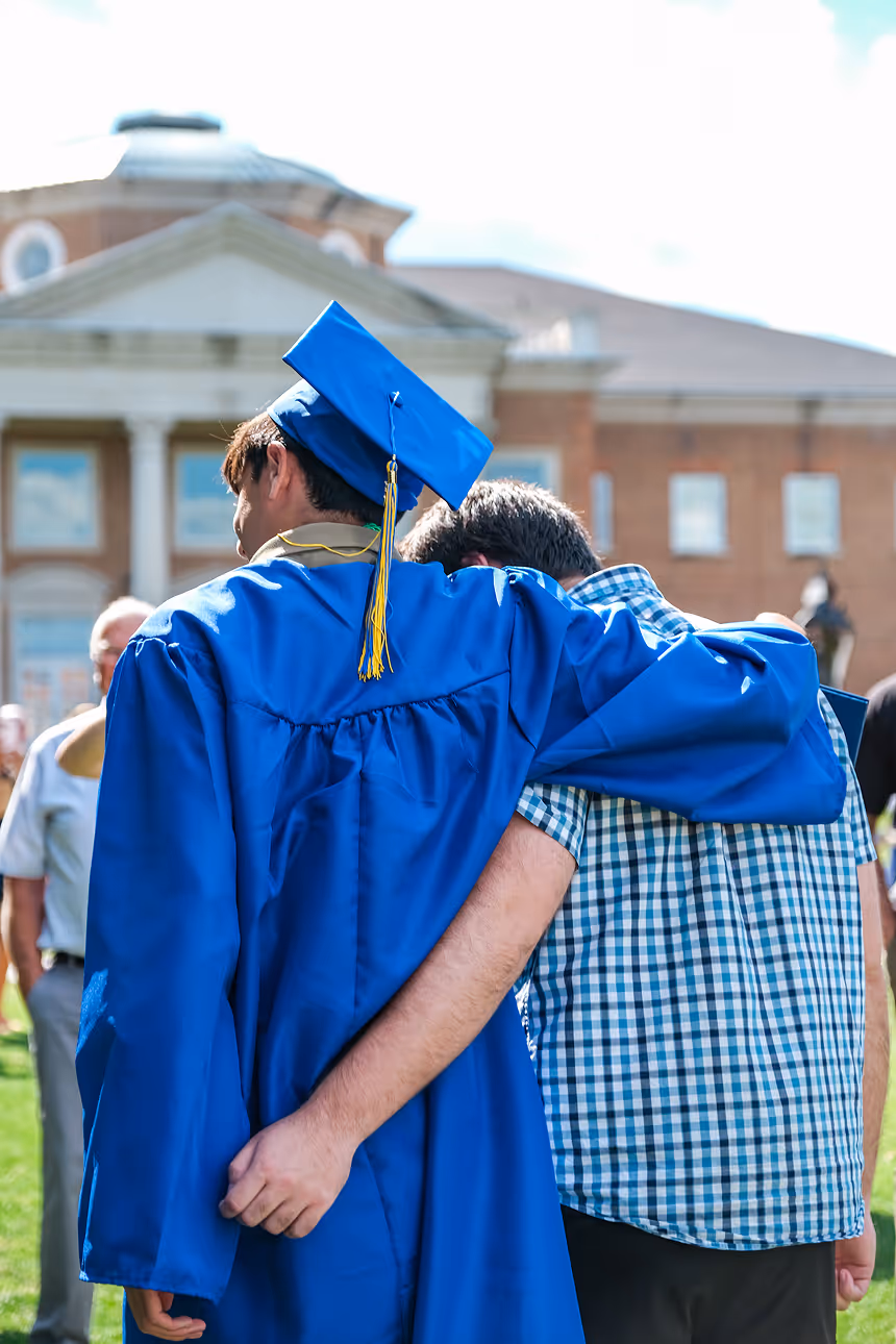 Cary Academy graduate in blue cap and gown embracing family member after graduation ceremony on campus.