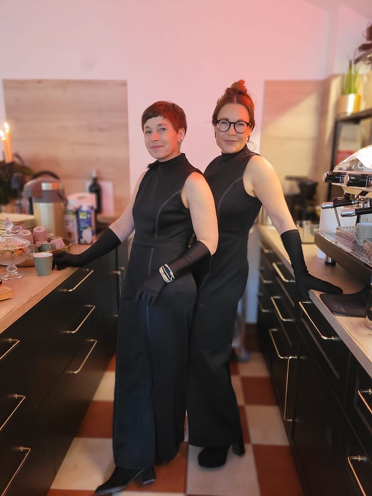 Two women dressed in matching black sleeveless outfits and gloves standing back-to-back in a kitchen or cafe setting.