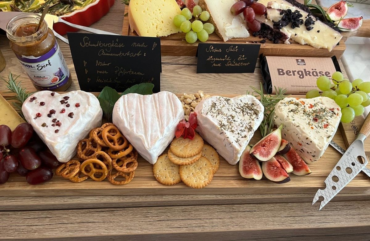 Wooden serving board with four heart-shaped cheeses garnished with herbs and peppercorns, accompanied by grapes, figs, crackers, pretzels, and a jar of mustard.