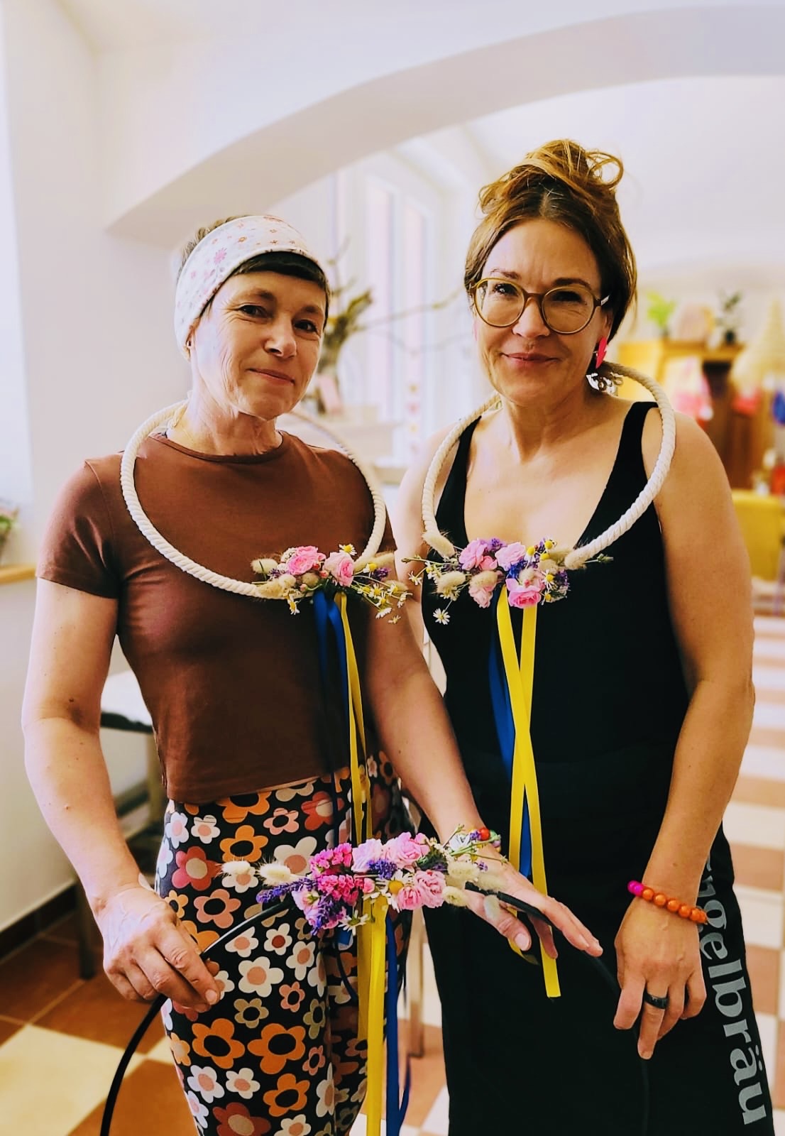 Two women indoors wearing floral necklaces with ribbons, one in a brown shirt and floral pants, the other in a black dress with glasses.