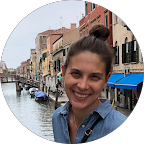Smiling woman with hair in a bun standing by a canal with boats and buildings in the background.