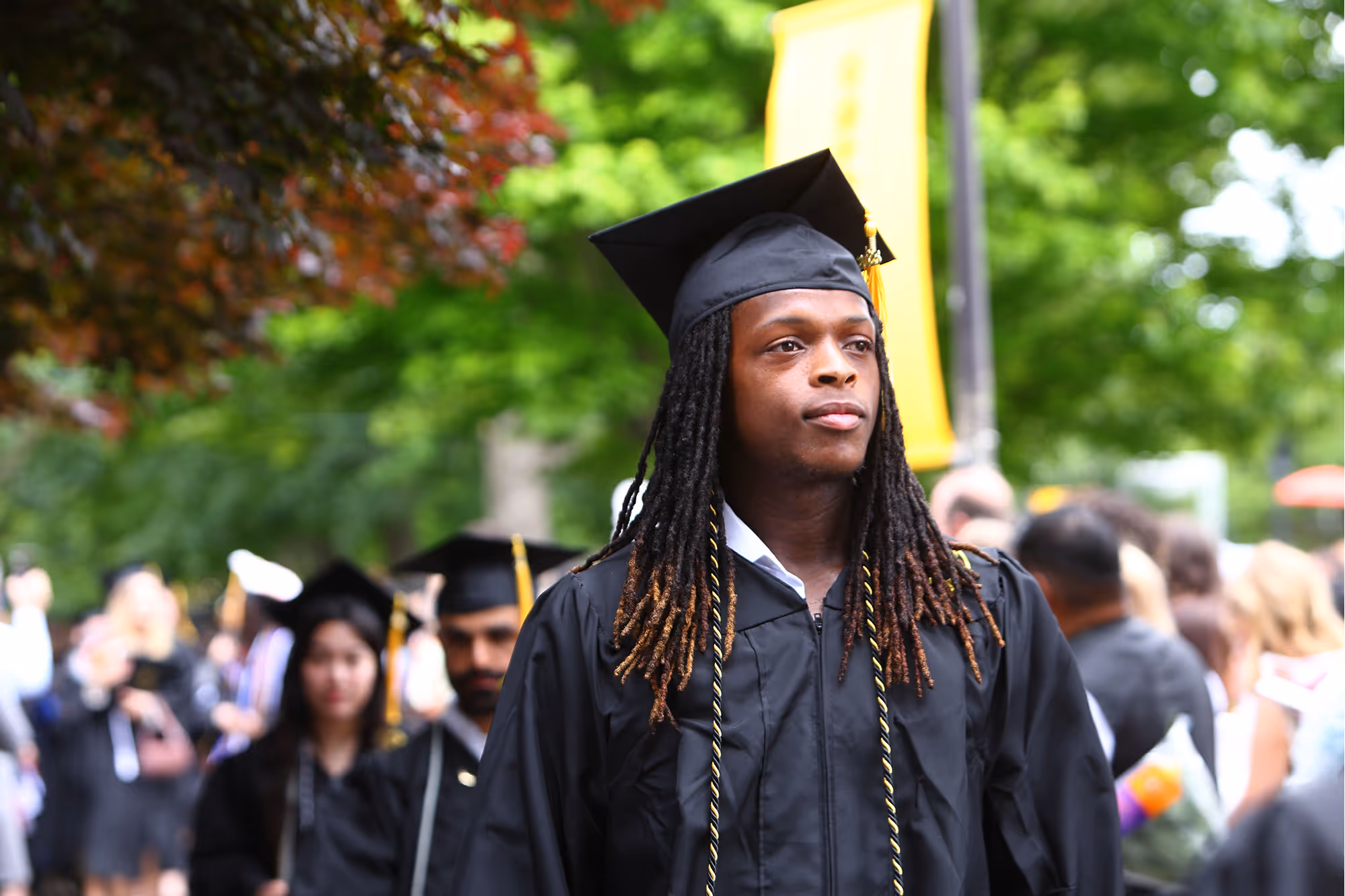 Male college student wearing graduation cap and gown, standing outdoors with blurred graduates and green trees in the background.