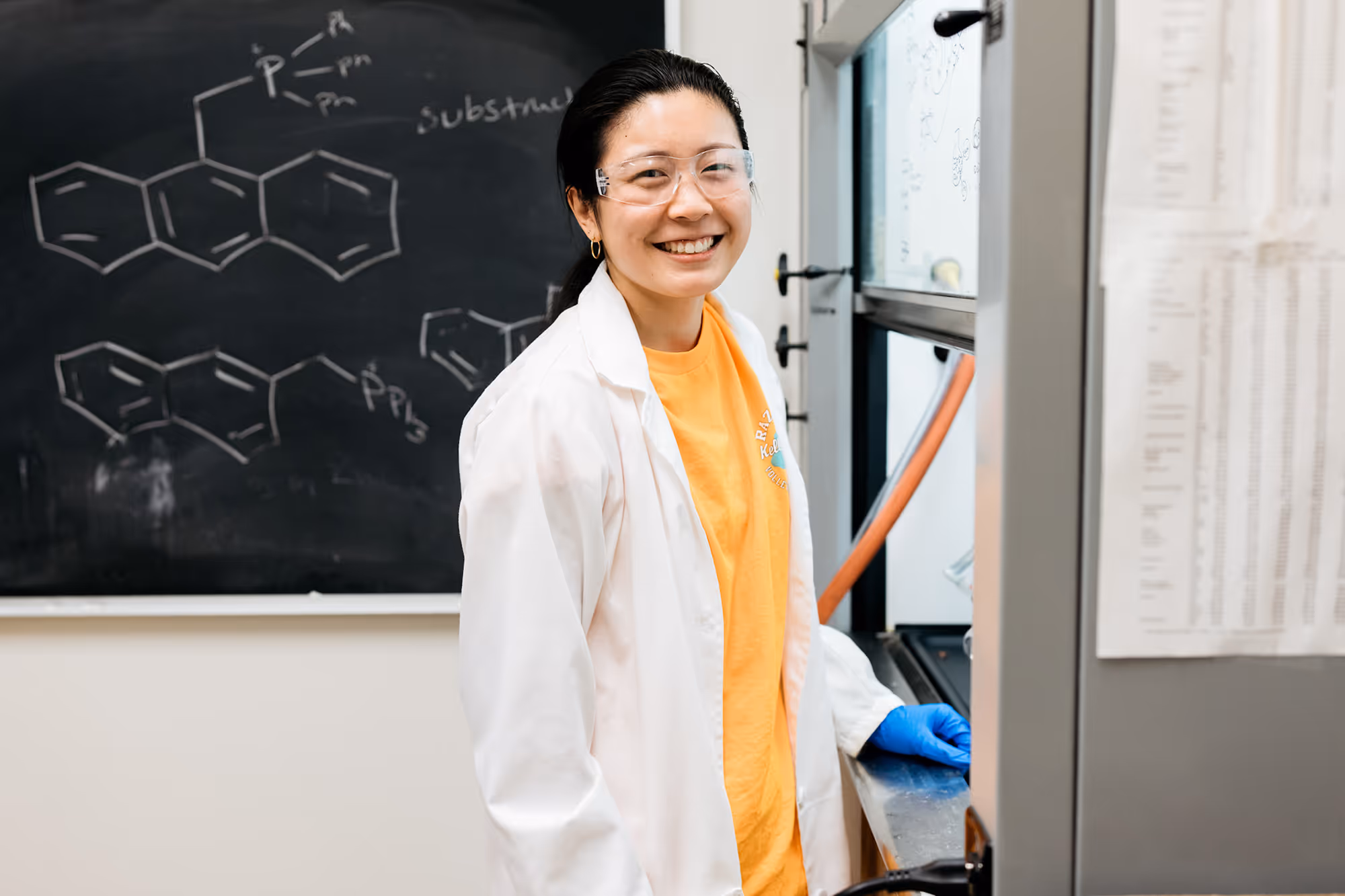 Smiling student scientist wearing safety glasses, a white lab coat, and blue gloves stands in a lab with chemical structures on a blackboard behind.