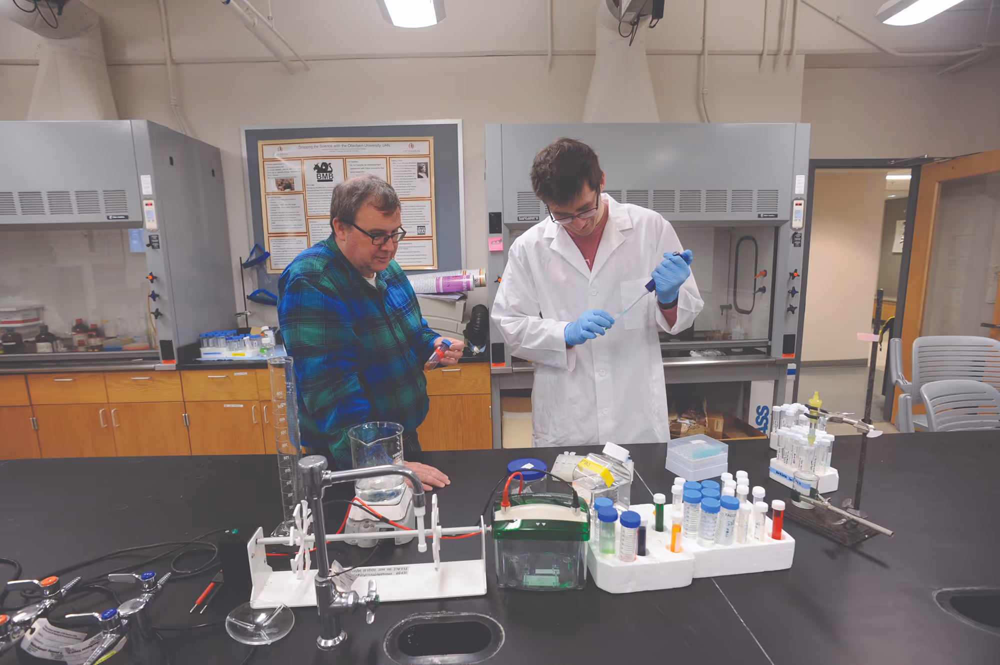 Two scientists in a lab, one in a white coat using a pipette, surrounded by lab equipment and test tubes.
