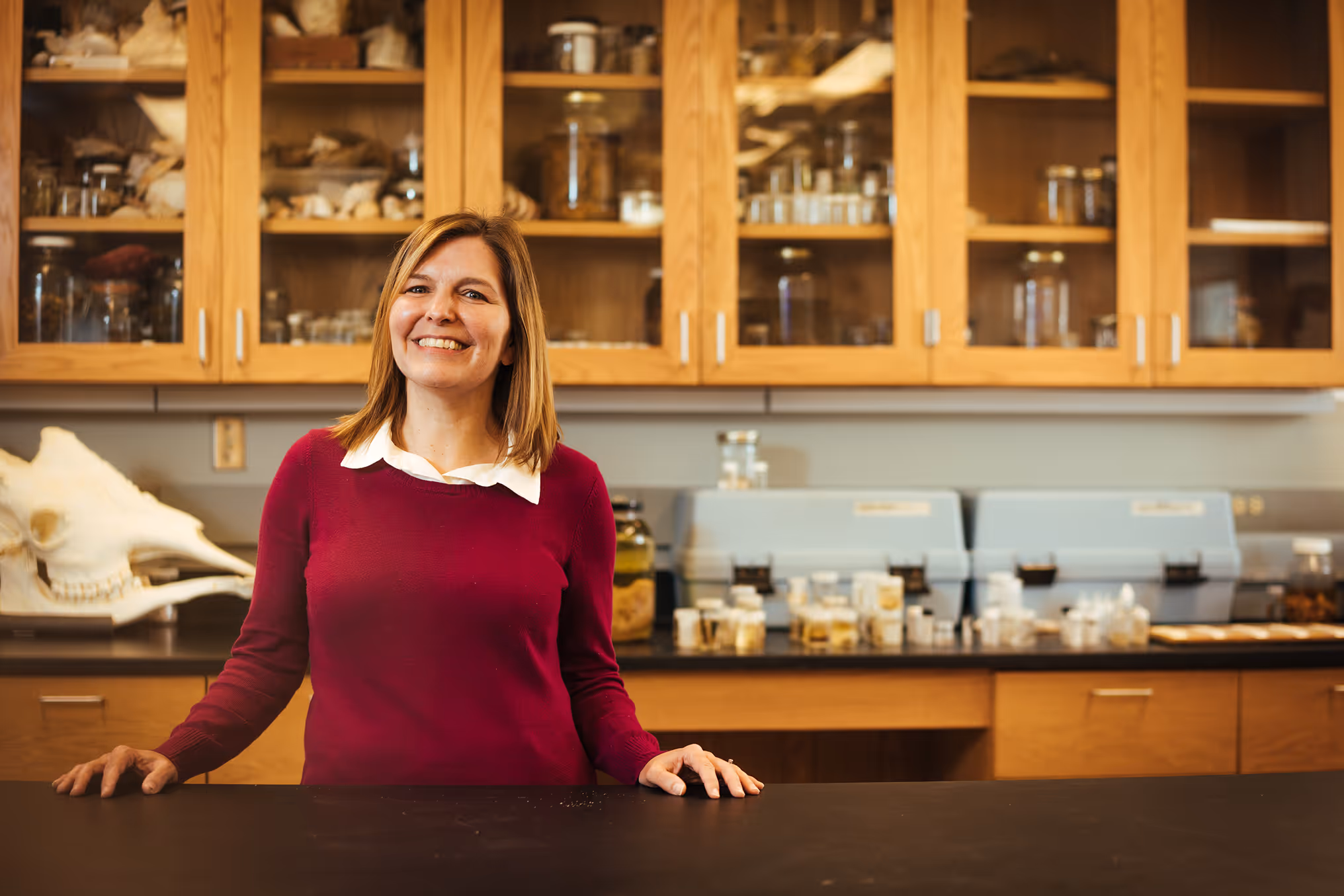 Smiling professor in a red sweater standing behind a lab counter with wooden cabinets and jars in the background.