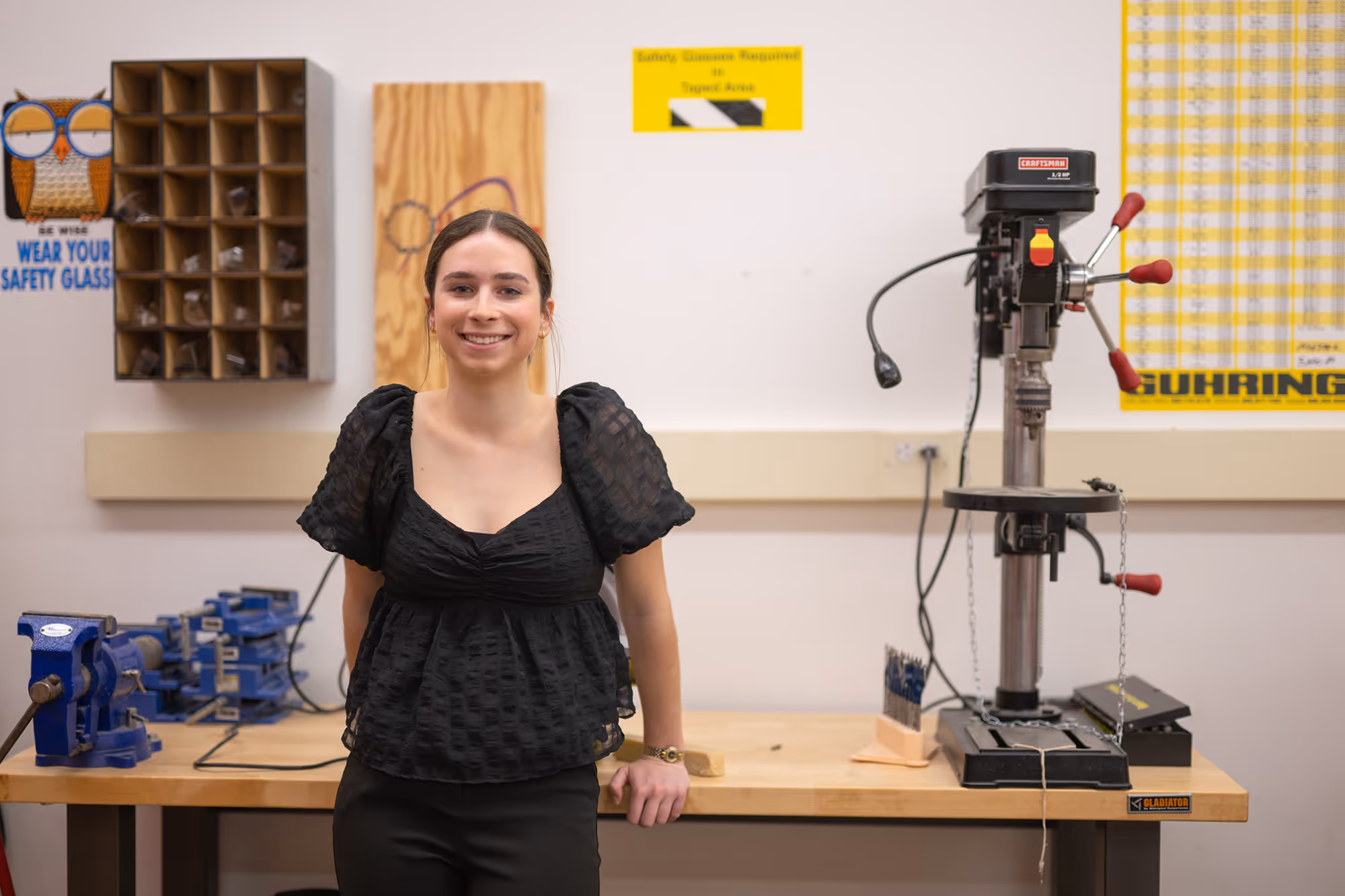 Smiling student leaning against a workbench with a drill press and blue vises in a workshop.
