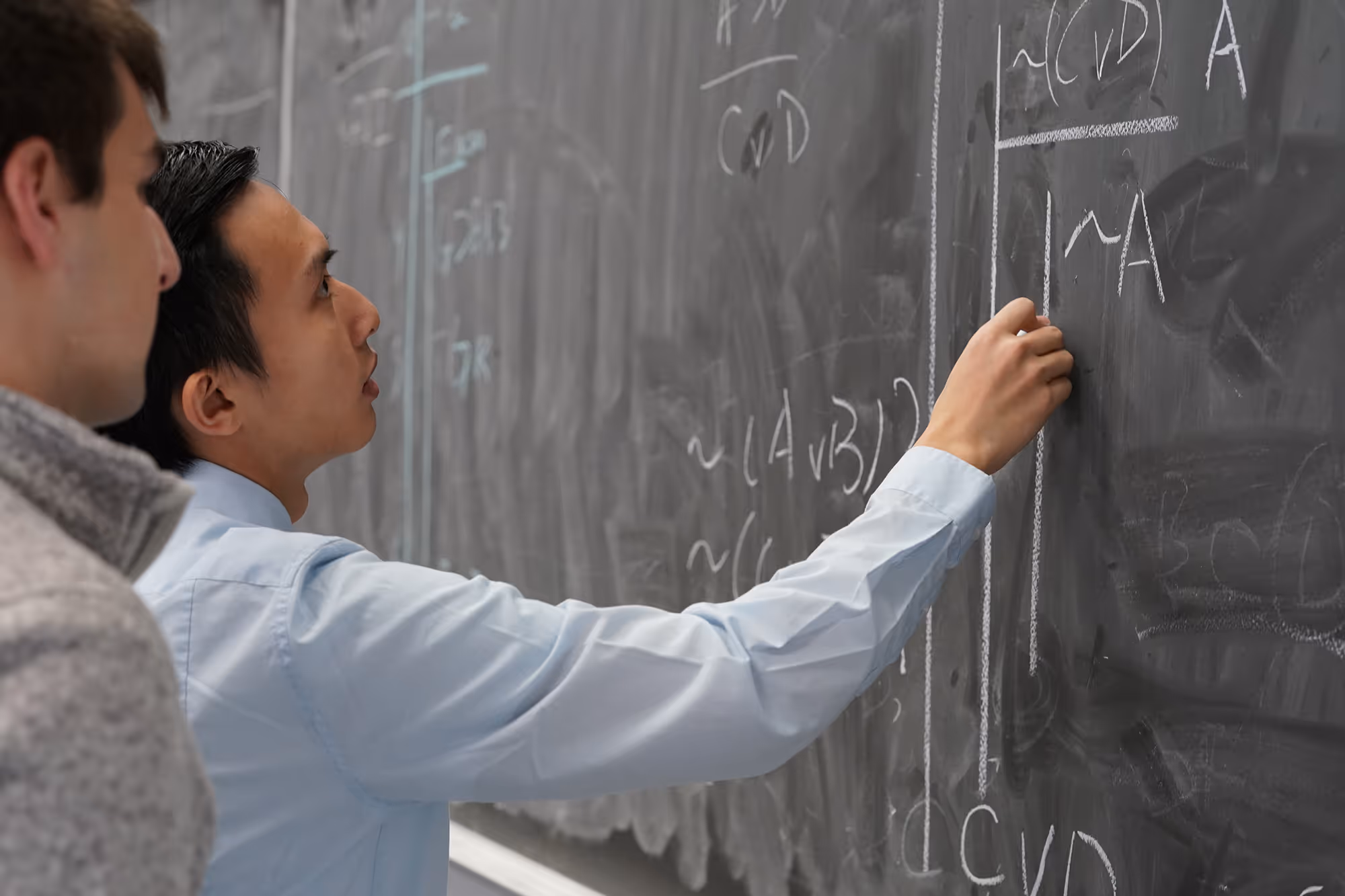 Two men at a chalkboard, one writing mathematical or logical formulas with chalk.
