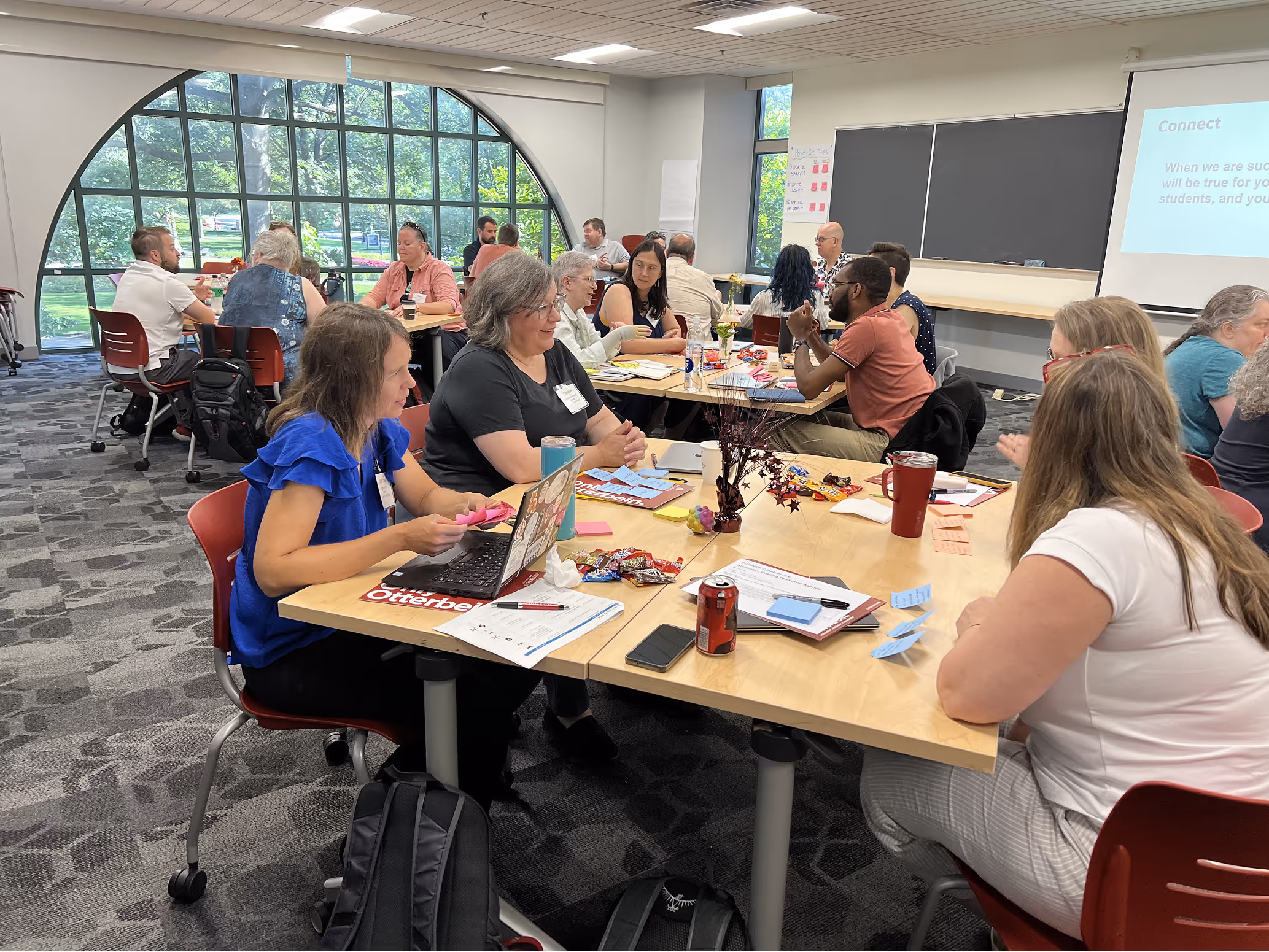 Group of people seated around tables in a classroom, discussing and working with laptops, notes, and snacks.