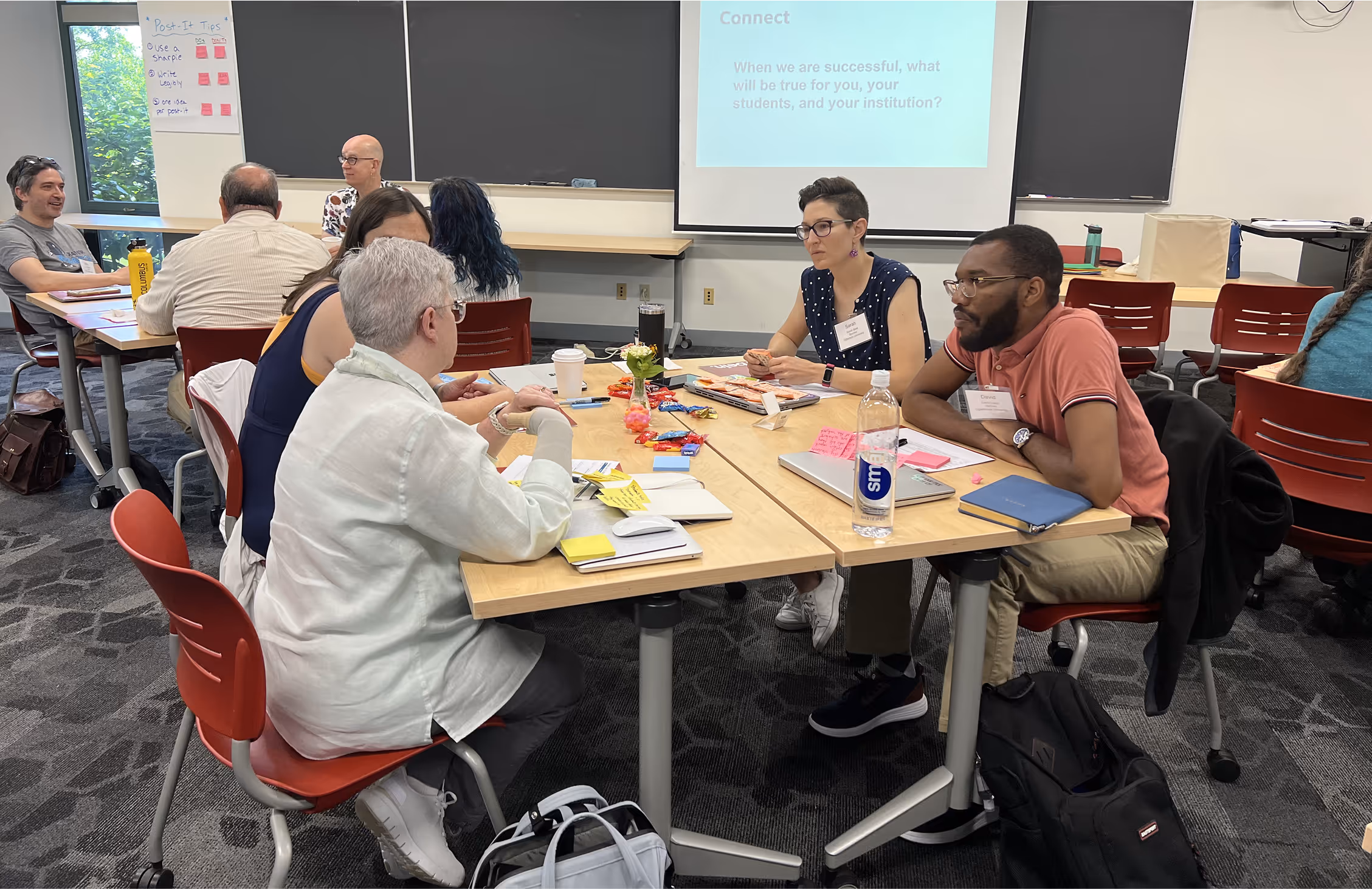 Group of adults seated around tables doing a workshop, engaging in discussion with notebooks, laptops, and snacks on the table.