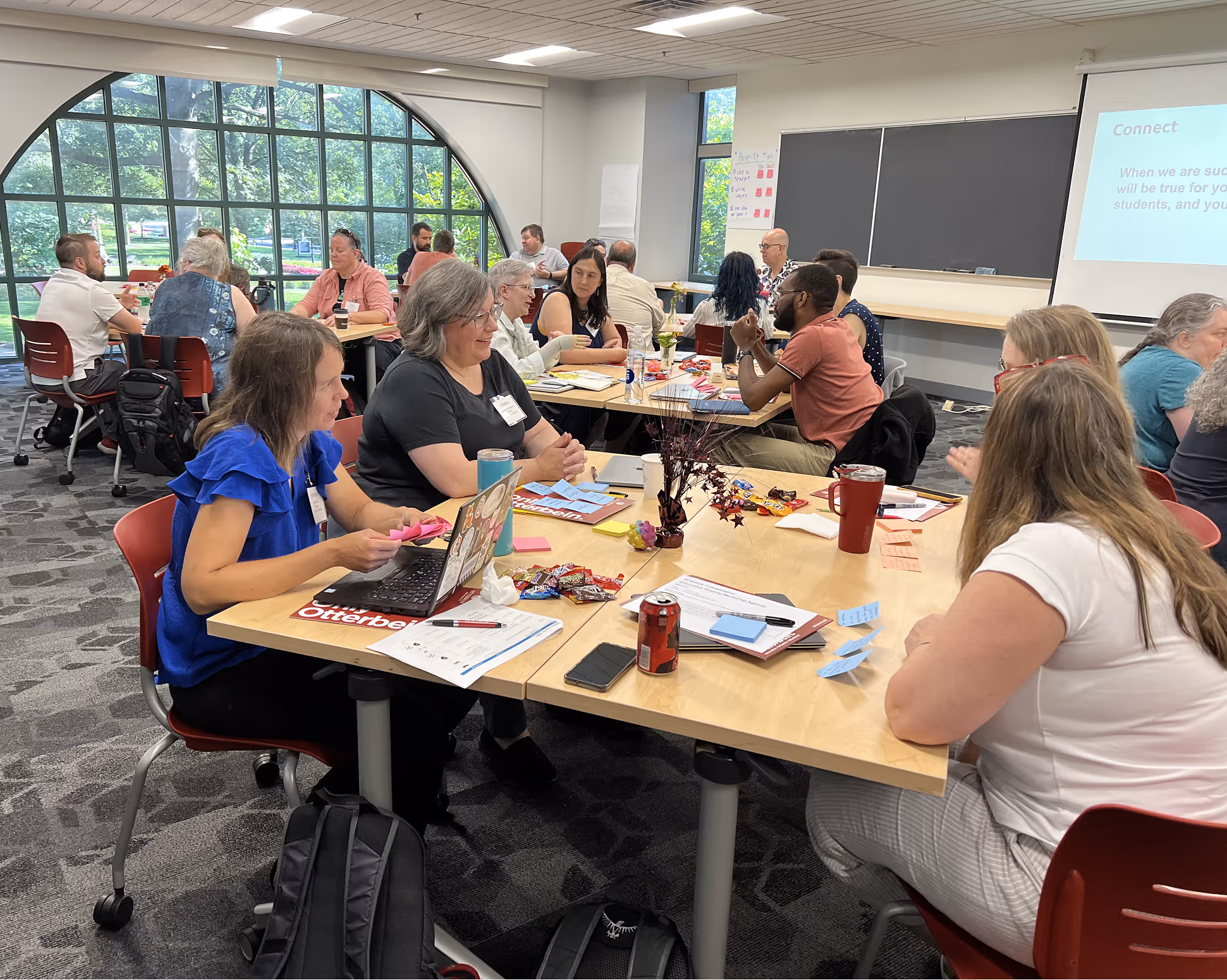 Group of adults seated at tables in a bright room with large window, engaged in a workshop or meeting with laptops, papers, and snacks.