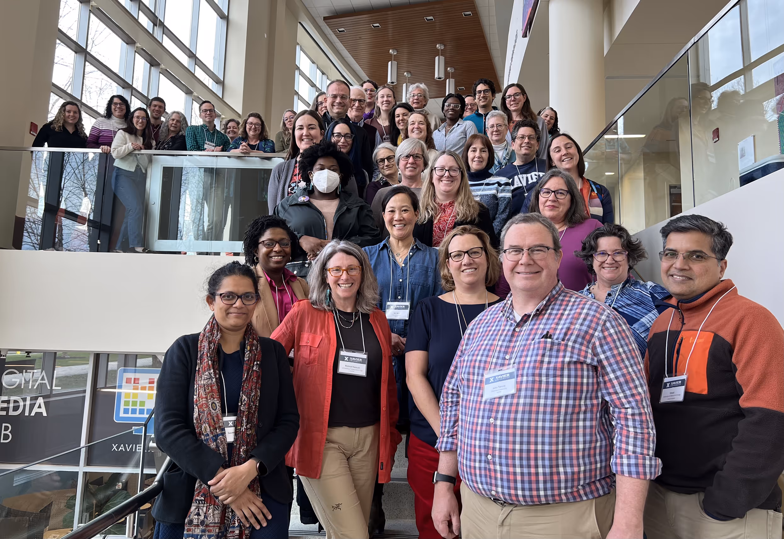 Group of staff and faculty smiling and standing on stairs inside a modern college building with large windows.