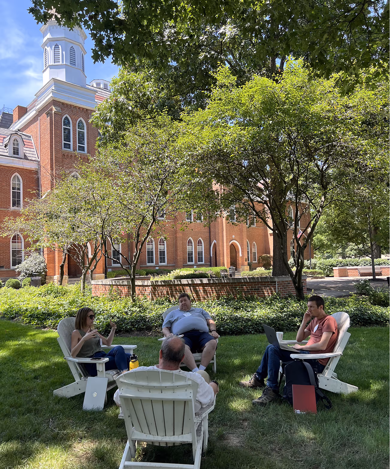 Four people seated in white Adirondack chairs on green lawn under trees in front of a red brick building on a sunny day.