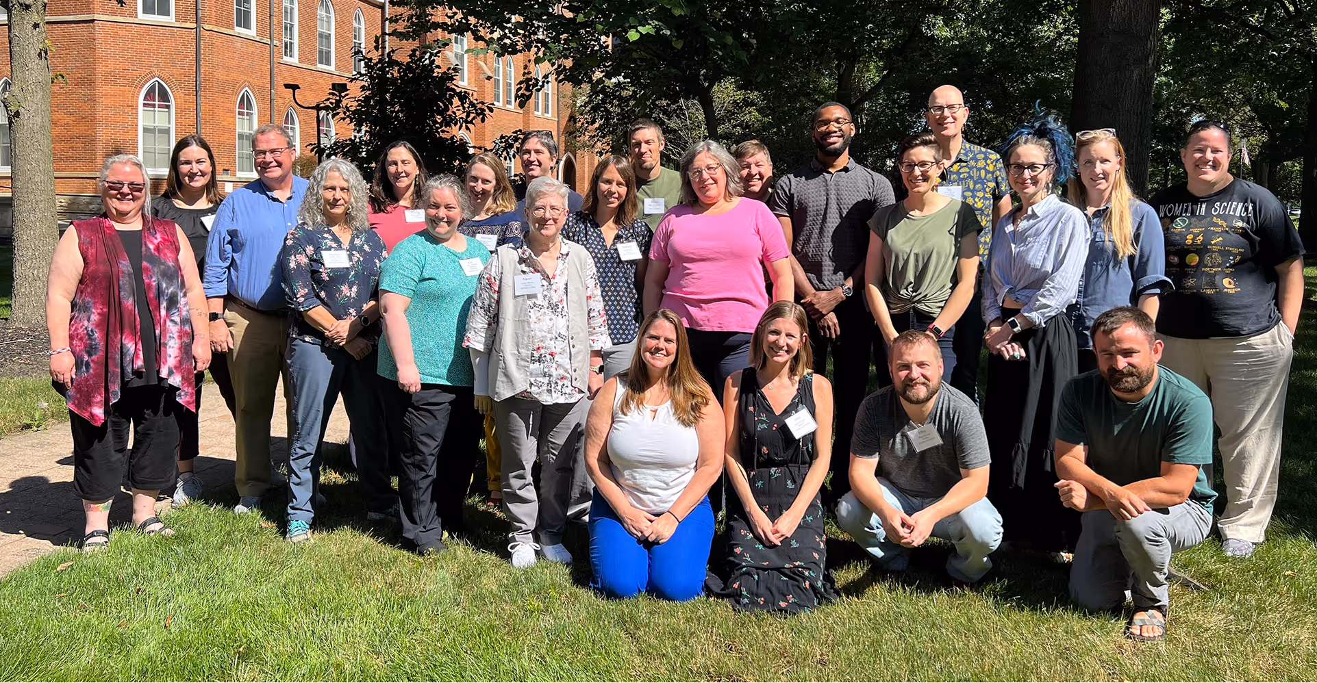 Group of 23 adults posing on green grass with a red brick building and shaded trees in the background on a sunny day.