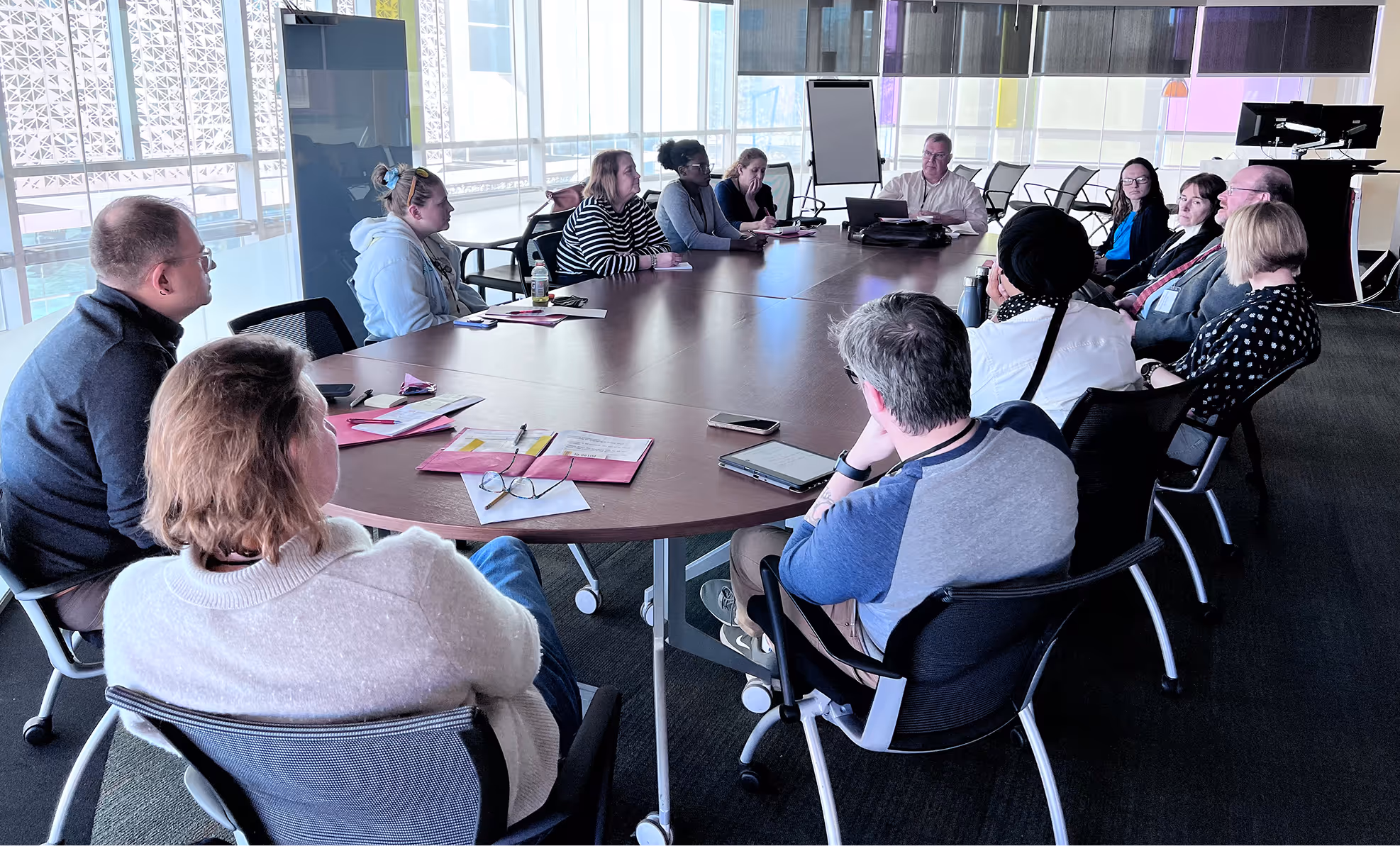 Group of twelve people seated around an oval conference table in a modern, sunlit meeting room.