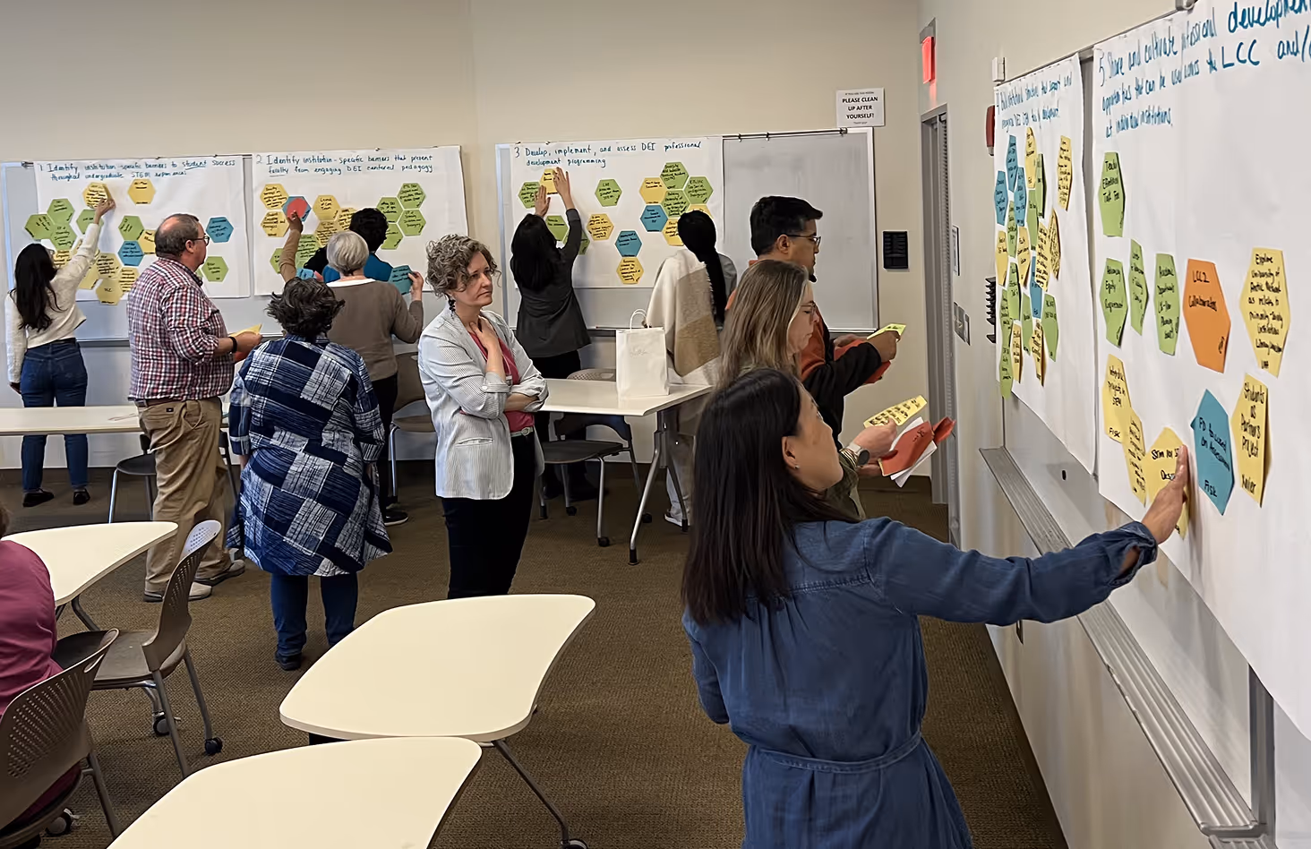 Group of people in a classroom placing colorful hexagonal sticky notes on large sheets of paper with handwritten text during a workshop.