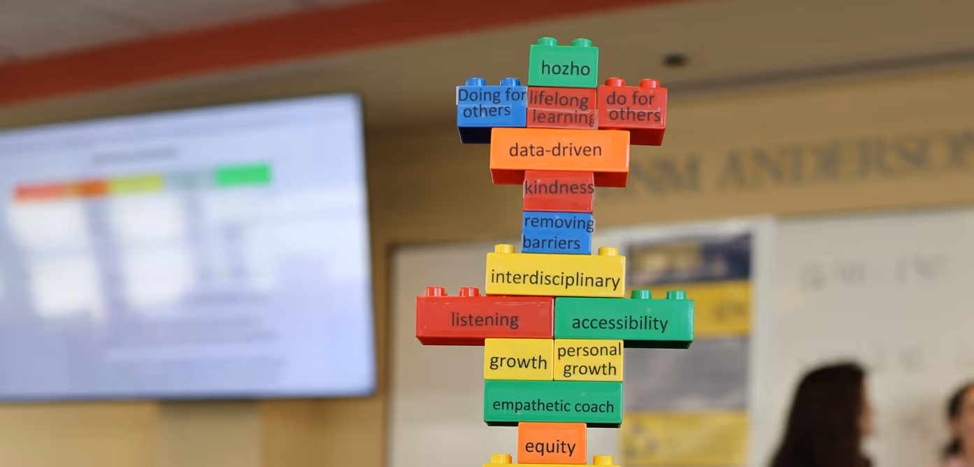 Stacked colorful blocks with words like 'data-driven,' 'interdisciplinary,' 'kindness,' and 'empathic coach' in a blurred classroom setting.