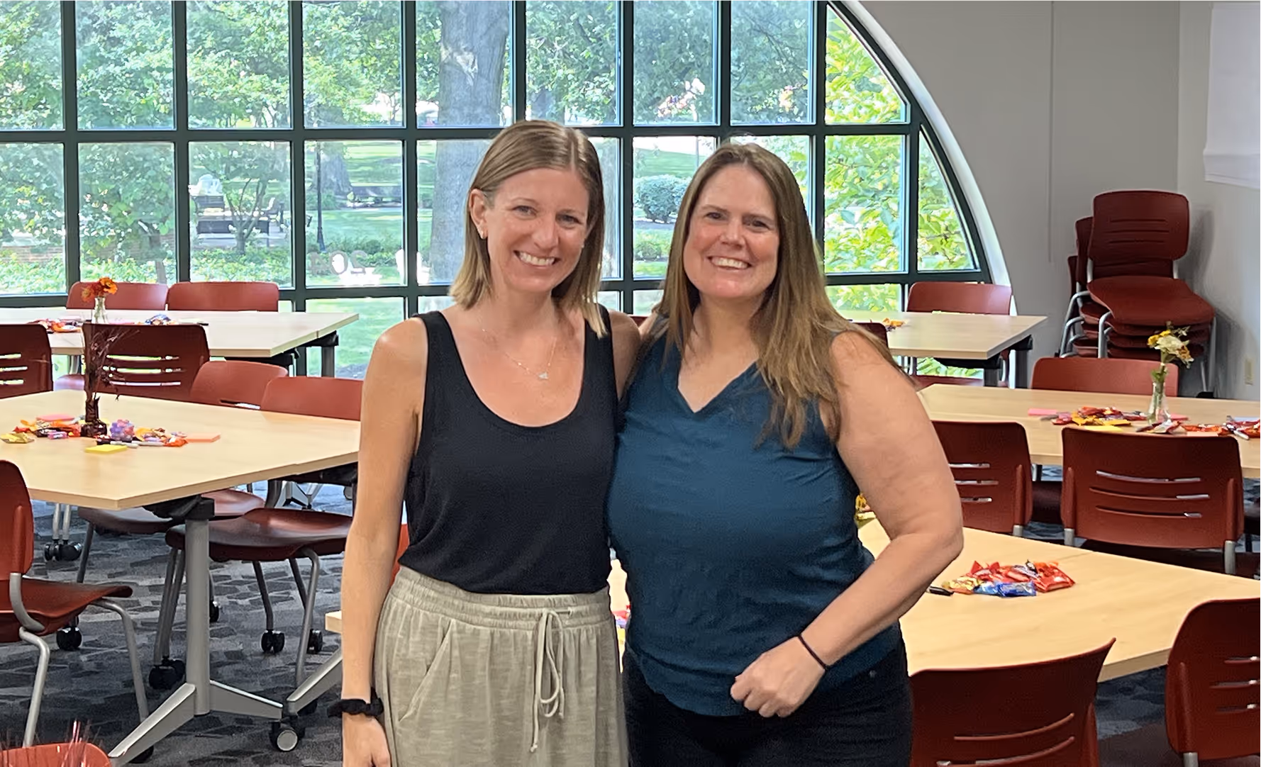 Two smiling women standing side by side in a bright room with large windows and tables with red chairs.