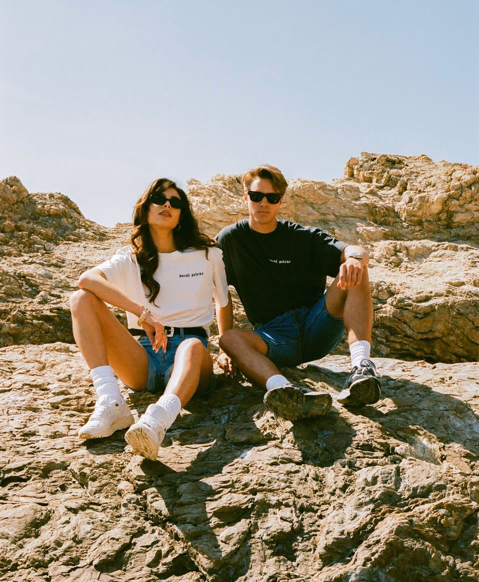 A man and a woman sitting on a rocky beach under a blue sky wearing Saint Selene t-shirt