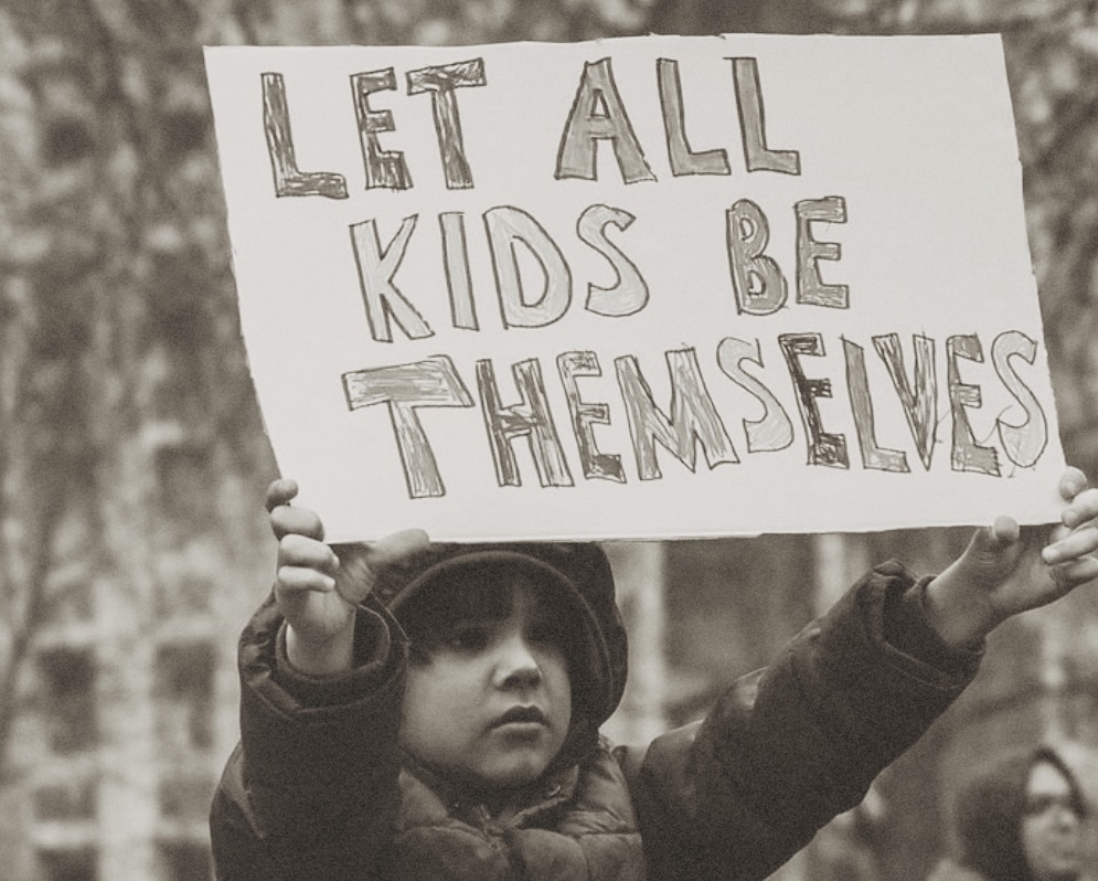 Young child wearing a winter coat and hat holding a sign that reads 'LET ALL KIDS BE THEMSELVES'.