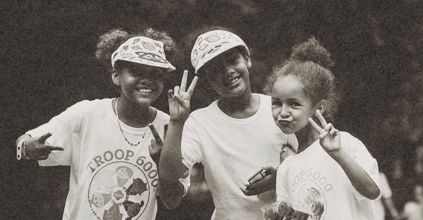 Three children smiling and posing with peace signs, wearing matching Troop 6000 t-shirts and hats.