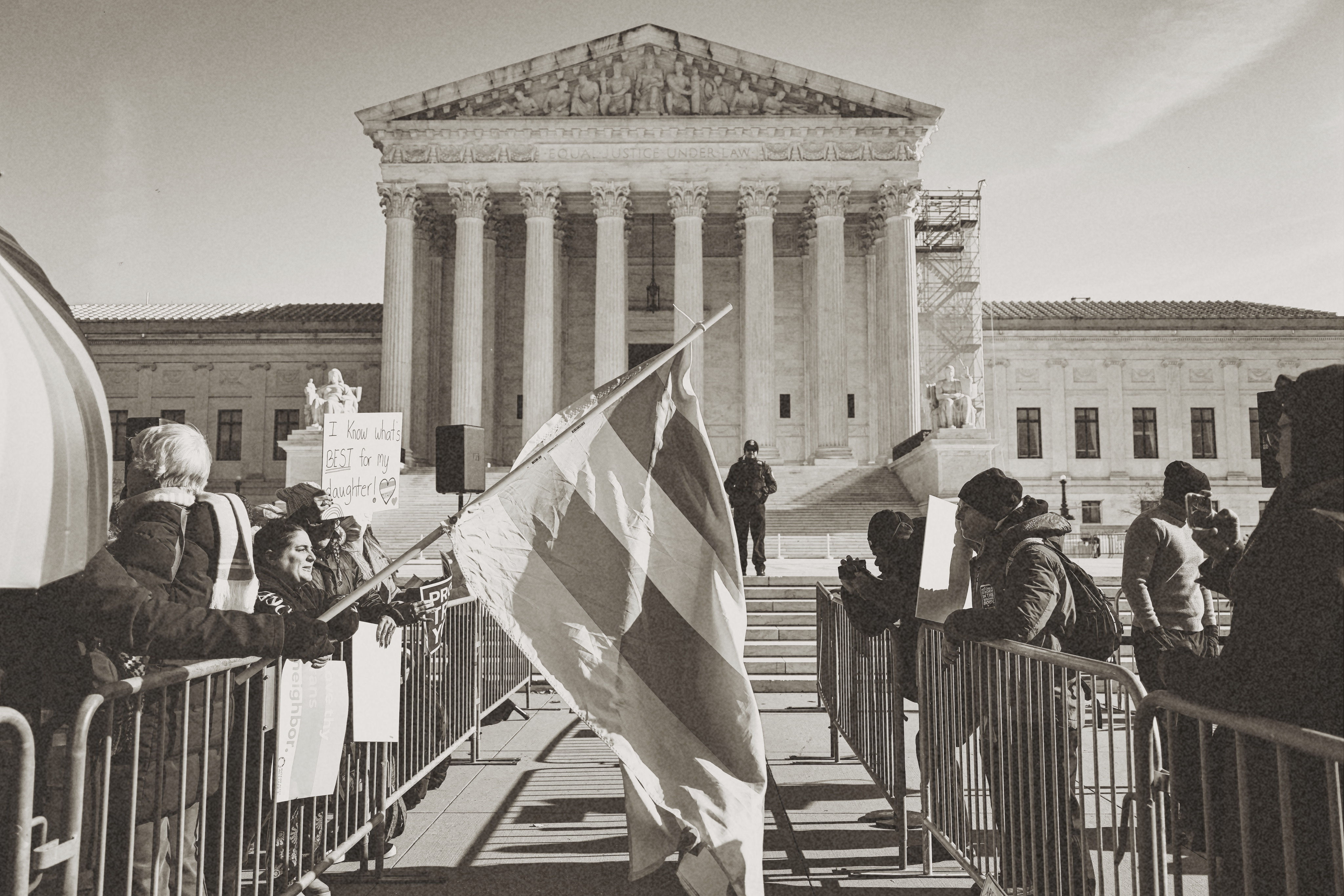 Protesters holding signs and a large flag outside the United States Supreme Court building with a police officer standing guard on the steps.