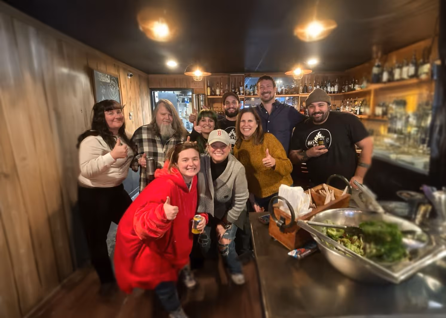 A group of people pose inside a cozy wood-paneled bar, smiling and giving thumbs up.