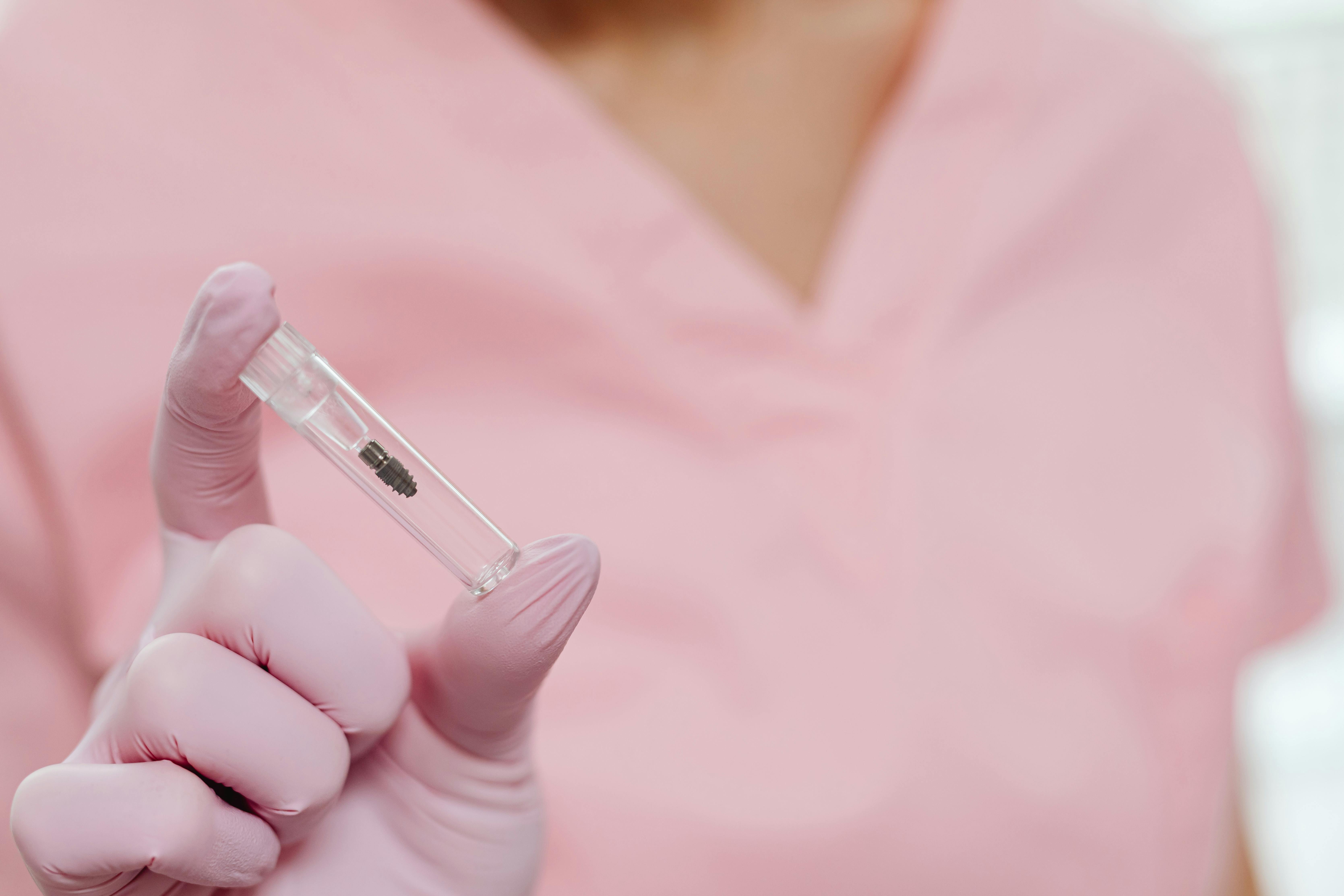 Gloved hand holding a small clear vial containing a tiny dental implant against a pink background.