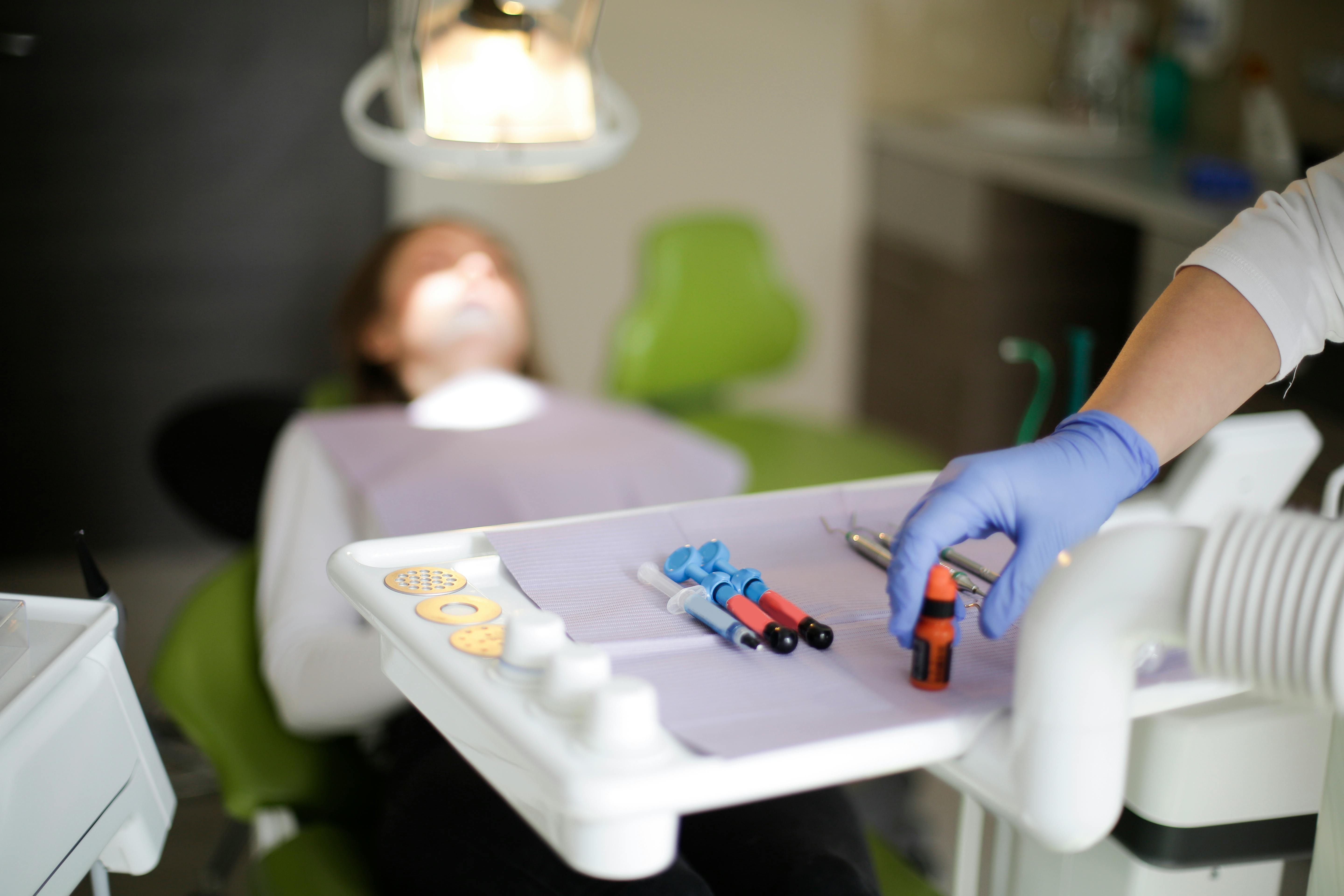 Dental tools and syringes on a tray with a gloved hand reaching for an item, in a dental clinic with a patient reclining in the background.