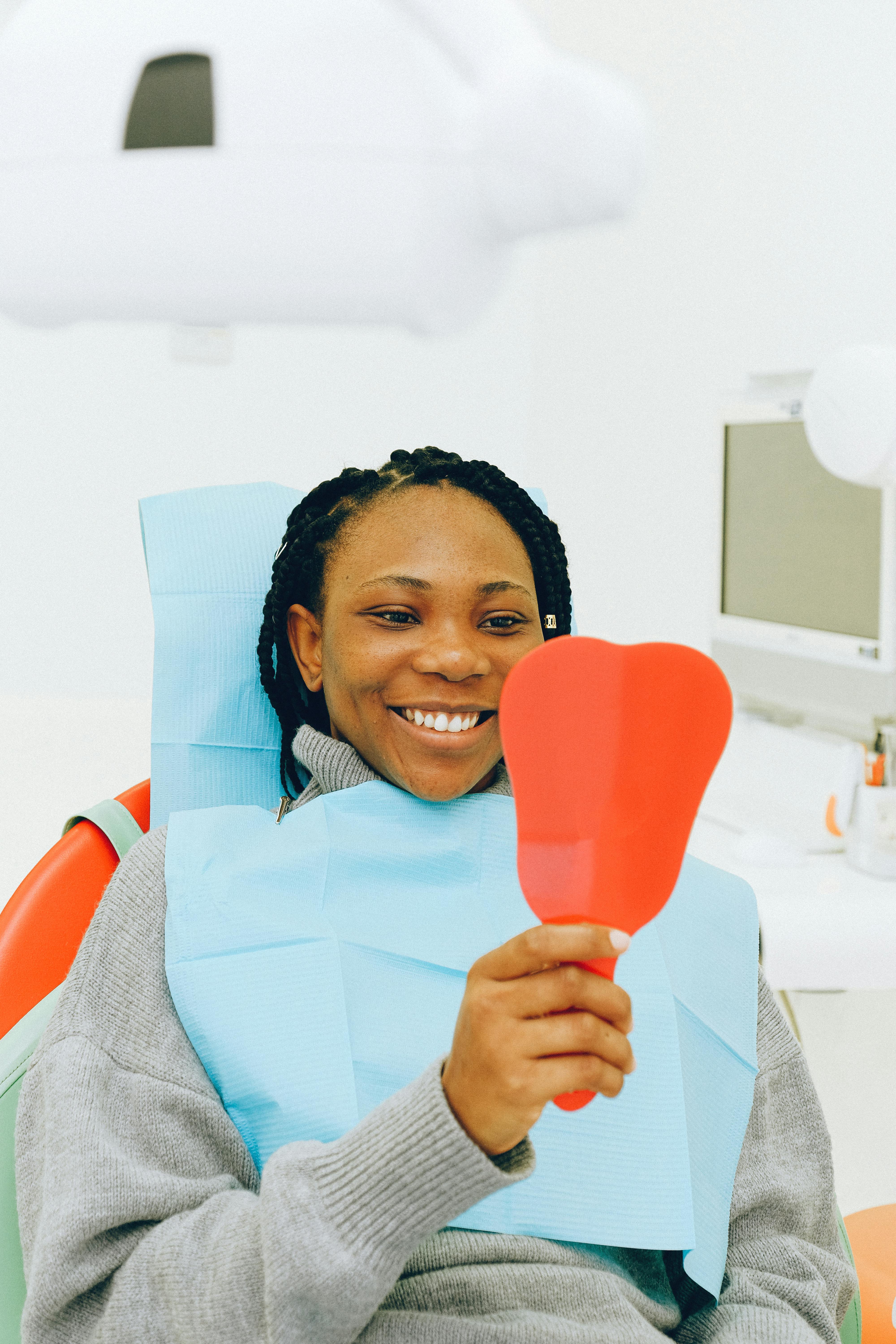 Smiling woman with braided hair sitting in a dental chair wearing a blue bib, holding a red mirror shaped like a tooth.