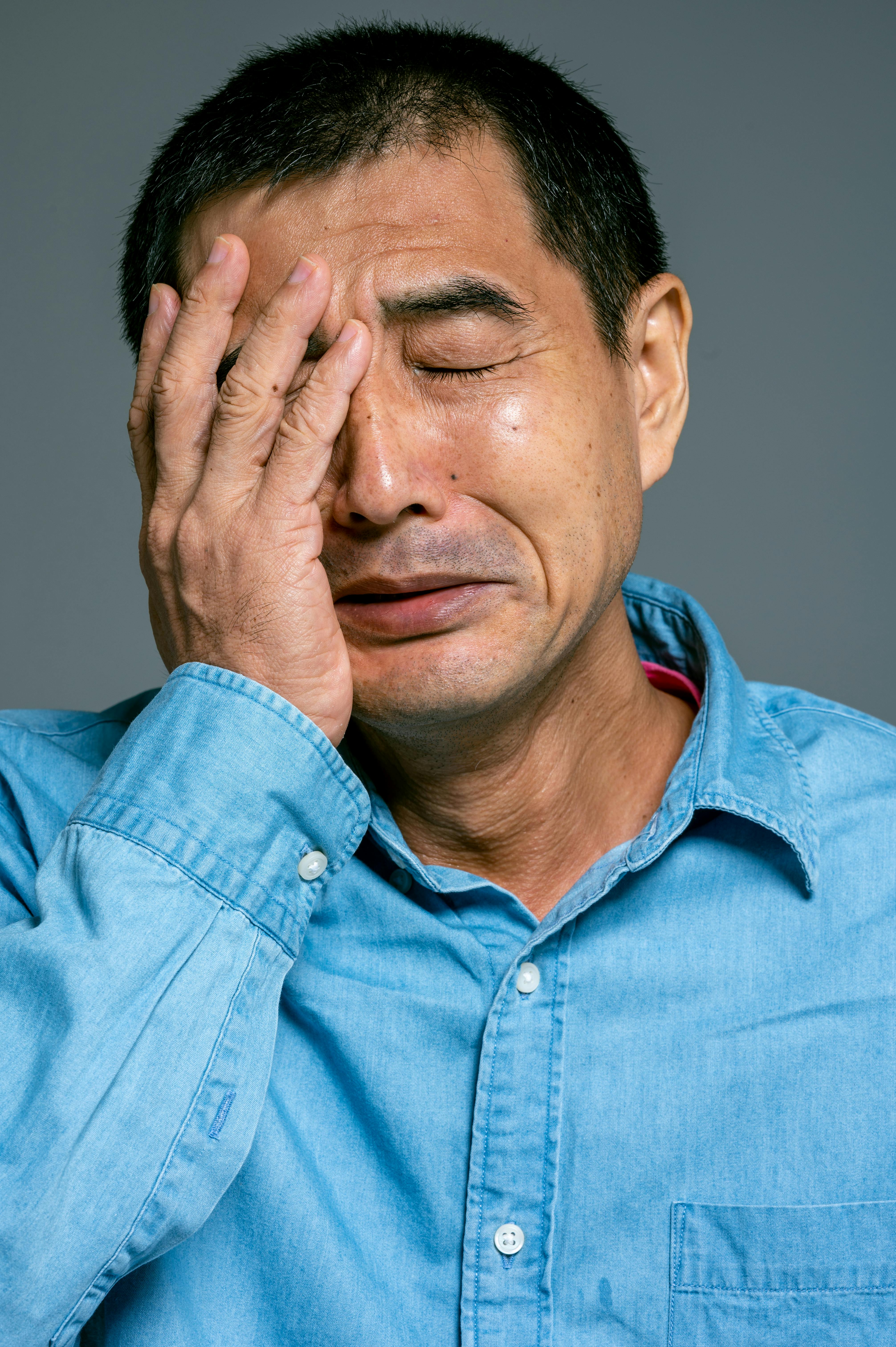 Middle-aged man in a light blue shirt with eyes closed, covering part of his face with his hand, appearing distressed.