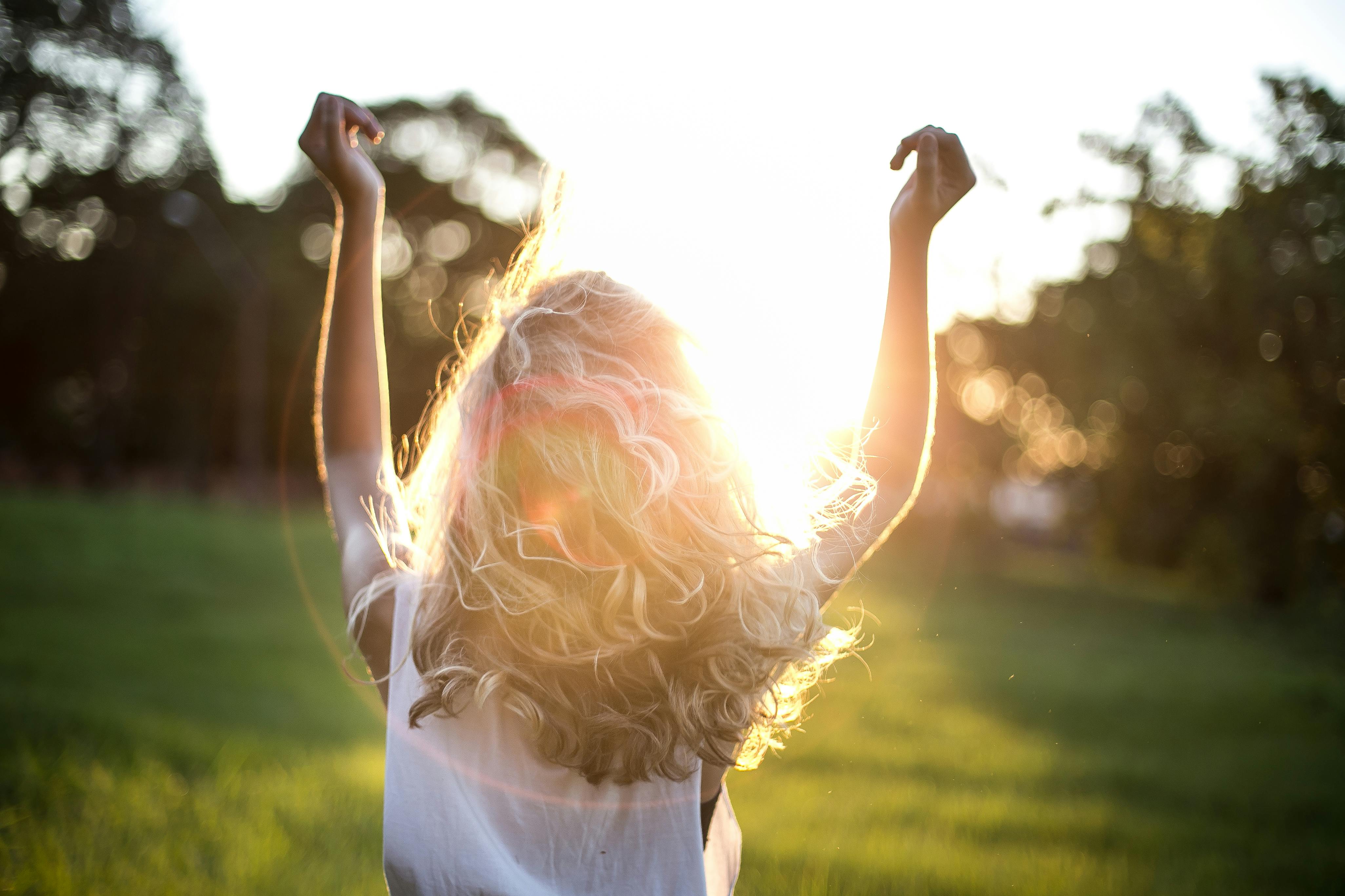 Person with curly blonde hair raising arms in a sunlit green field during sunset.